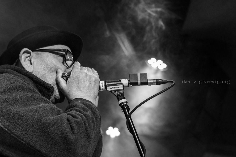 Primer plano lateral del armonicista Lalo, con sombrero y gafas, pegado al micrófono mientras sopla su armónica. De fondo se ve el humo mezclándose con el humo de la sala Groove.