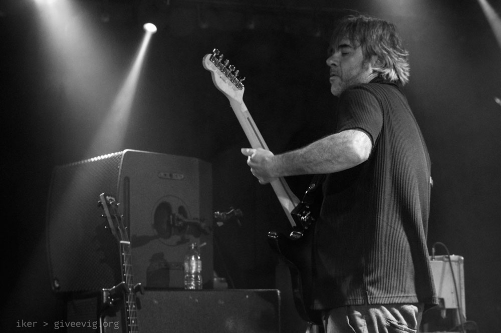 Iñaki, casi de espaldas al público pero fotografiado desde el lado izquierdo, levanta sutilmente su guitarra mientras realiza un punteo.