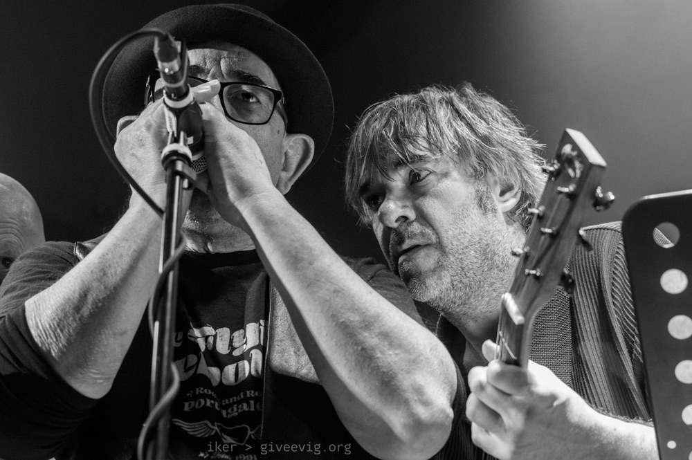 Primer plano frontal de Lalo mientras sopla su armónica pegada al micrófono y por la derecha se le arrima Iñaki.