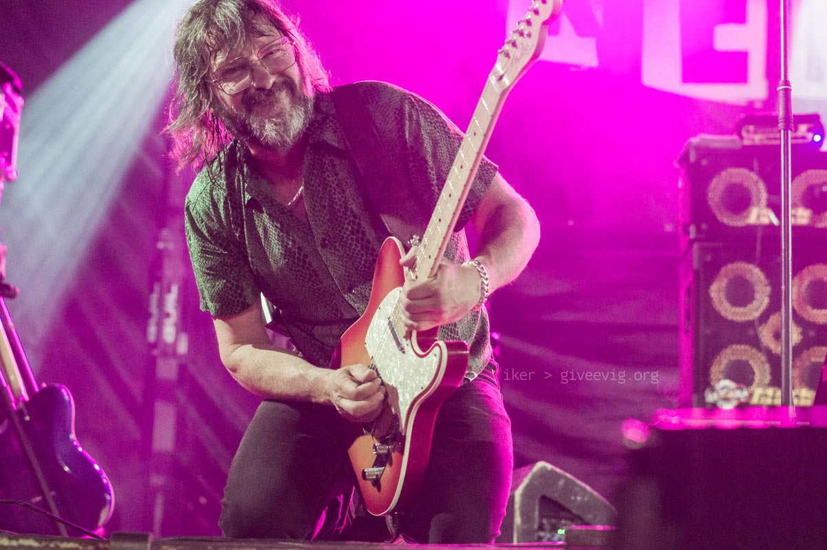 Gorka, de rodillas y con cara de esfuerzo, tocando la guitarra bajo focos lilas.