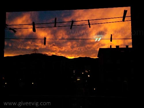 Amanecer naranja con nubes moradas, con las siluetas de unas cuerdas con pinzas y edificios y calles oscuras